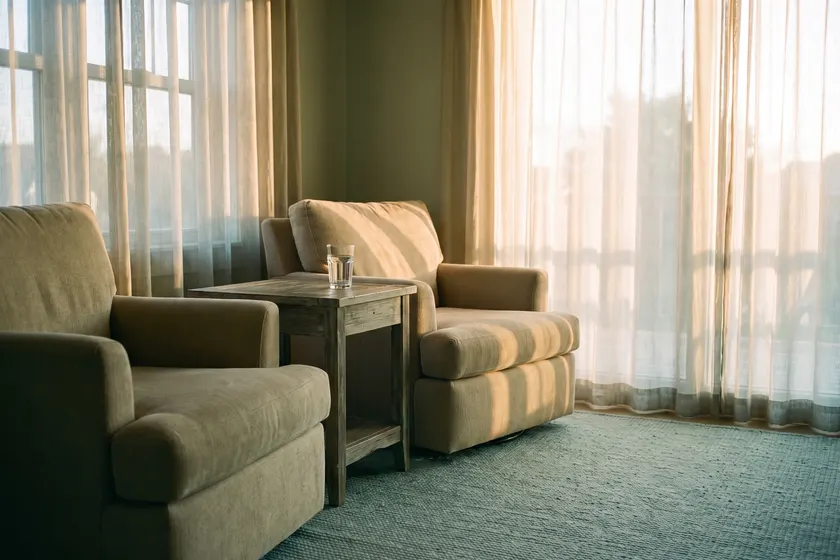 Two armchairs angled toward each other by a sunlit window with a glass of water on a side table